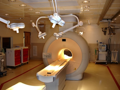 A hospital room containing an MRI machine surrounded by overhead lights and various medical equipment. The room has a clean and clinical environment with red cabinets on the left and diagnostic machines on the right.