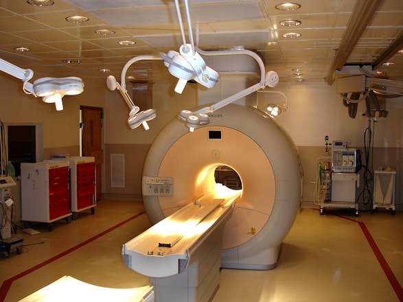 A hospital room containing an MRI machine surrounded by overhead lights and various medical equipment. The room has a clean and clinical environment with red cabinets on the left and diagnostic machines on the right.