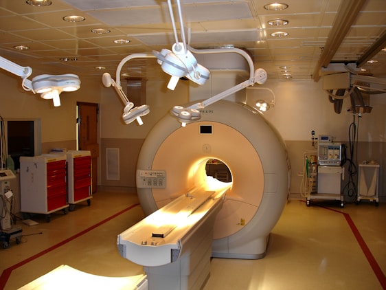 A hospital room containing an MRI machine surrounded by overhead lights and various medical equipment. The room has a clean and clinical environment with red cabinets on the left and diagnostic machines on the right.