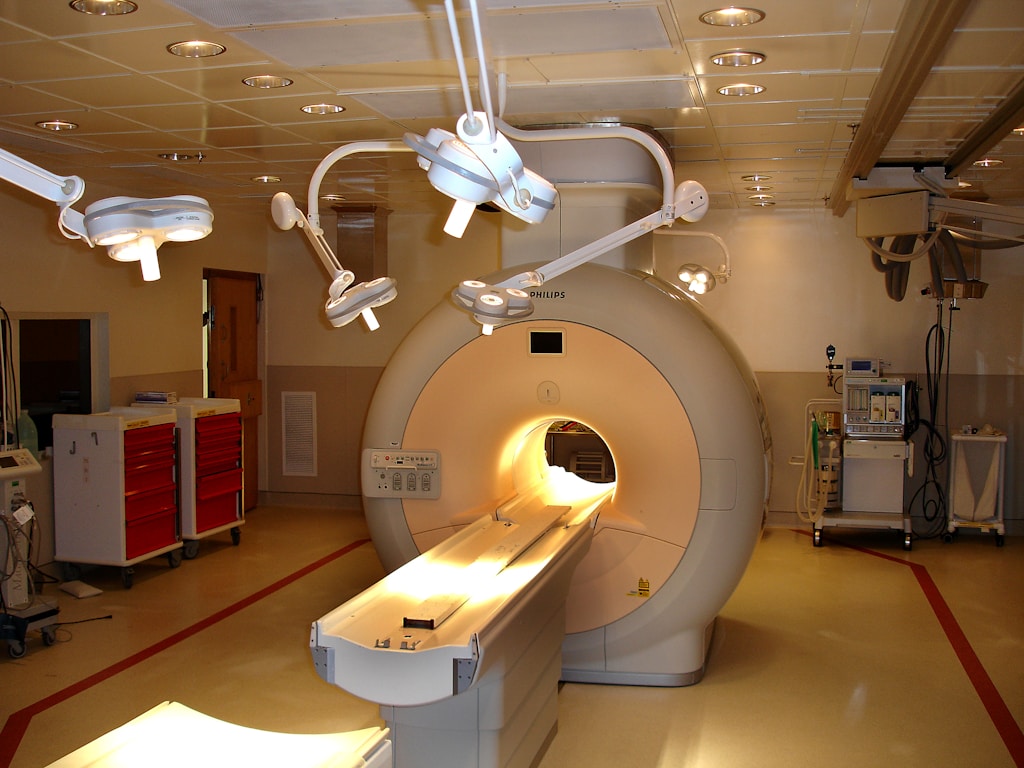 A hospital room containing an MRI machine surrounded by overhead lights and various medical equipment. The room has a clean and clinical environment with red cabinets on the left and diagnostic machines on the right.
