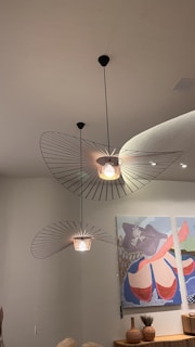 Stylish pendant lights with geometric metal shades hang from the ceiling. Beneath them, a modern art piece with abstract shapes and vibrant colors is displayed on the wall. A wooden shelf holds decorative vases and a dry floral arrangement.