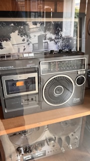 A vintage radio cassette recorder is displayed on a wooden shelf. The device features a large speaker, multiple dials, and a well-preserved cassette slot. The design is reminiscent of technology from the past, with its metallic accents and analog display. Behind the glass, reflection of trees and a balcony is visible, adding depth to the scene.