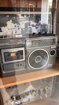 A vintage radio cassette recorder is displayed on a wooden shelf. The device features a large speaker, multiple dials, and a well-preserved cassette slot. The design is reminiscent of technology from the past, with its metallic accents and analog display. Behind the glass, reflection of trees and a balcony is visible, adding depth to the scene.