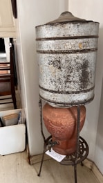 A rustic, vintage water dispenser is composed of a large, weathered metal container atop a terracotta base. The metal container appears old and slightly corroded, with visible signs of wear. The terracotta base features decorative elements and has a tap attached, resting on a metallic stand with intricate design.