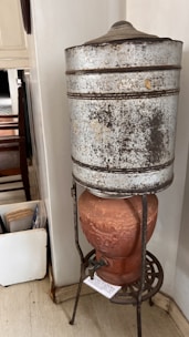 A rustic, vintage water dispenser is composed of a large, weathered metal container atop a terracotta base. The metal container appears old and slightly corroded, with visible signs of wear. The terracotta base features decorative elements and has a tap attached, resting on a metallic stand with intricate design.