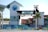 An outdoor venue with a stage that has a sign reading 'Hangout'. There are numerous blue and turquoise chairs and tables arranged in front of the stage. The area is decorated with palm trees and string lights. In the background, there's a large building with a logo for 'Grand Strand Coastal Realty'. The scene appears to be set up for a casual gathering or event.