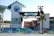 An outdoor venue with a stage that has a sign reading 'Hangout'. There are numerous blue and turquoise chairs and tables arranged in front of the stage. The area is decorated with palm trees and string lights. In the background, there's a large building with a logo for 'Grand Strand Coastal Realty'. The scene appears to be set up for a casual gathering or event.
