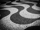 A monochrome patterned pavement composed of interlocking stone tiles forming undulating wave-like designs. The alternating dark and light stones create a striking visual effect with smooth, flowing lines.