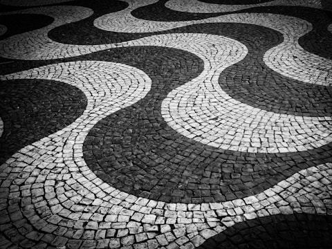 A monochrome patterned pavement composed of interlocking stone tiles forming undulating wave-like designs. The alternating dark and light stones create a striking visual effect with smooth, flowing lines.