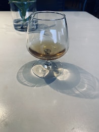 A snifter glass with a small amount of amber liquid sits on a light-colored table, casting soft shadows. In the background, there is a blurred glass vase with a green plant.