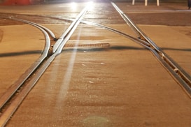 Two railway tracks intersect on a paved surface under a warm, ambient light at night. The converging tracks create a unique pattern, with light reflections adding depth and texture to the scene. The glow from a distant light source enhances the atmospheric quality.