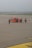 An orange airport vehicle is positioned on a large tarmac, accompanied by several workers wearing safety vests. The background includes a runway and distant airport infrastructure, all under an overcast sky.