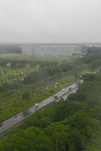 A large industrial building with a modern design is situated in the background, surrounded by a well-maintained grassy area with planted trees. In the foreground, a road with several vehicles of different sizes is visible, running parallel to a set of railway tracks. The scene is enveloped in a cloudy and misty atmosphere, suggesting recent or ongoing rainfall.