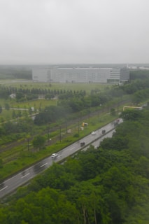 A large industrial building with a modern design is situated in the background, surrounded by a well-maintained grassy area with planted trees. In the foreground, a road with several vehicles of different sizes is visible, running parallel to a set of railway tracks. The scene is enveloped in a cloudy and misty atmosphere, suggesting recent or ongoing rainfall.