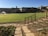 A well-maintained sports field is bordered by a stone pathway and decorative railing in the foreground, with large buildings in the background. A water sprinkler is active on the field, and two small soccer goals are visible at the far end. The scene is illuminated by bright sunlight, casting clear shadows.
