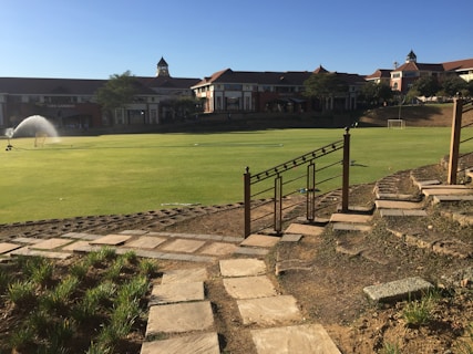 A well-maintained sports field is bordered by a stone pathway and decorative railing in the foreground, with large buildings in the background. A water sprinkler is active on the field, and two small soccer goals are visible at the far end. The scene is illuminated by bright sunlight, casting clear shadows.