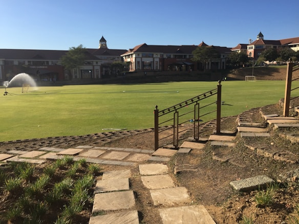 A well-maintained sports field is bordered by a stone pathway and decorative railing in the foreground, with large buildings in the background. A water sprinkler is active on the field, and two small soccer goals are visible at the far end. The scene is illuminated by bright sunlight, casting clear shadows.