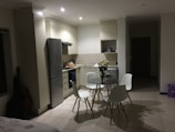 A modern kitchen with a minimalist design features white cabinets, a stainless steel refrigerator, and a small dining area with a round glass table and four white chairs. A vase of white flowers is placed on the table, adding a touch of elegance. The lighting is soft, creating a peaceful ambiance. There's a guitar leaning against the wall, and in the background, various kitchen items are neatly organized.