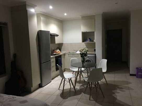 A modern kitchen with a minimalist design features white cabinets, a stainless steel refrigerator, and a small dining area with a round glass table and four white chairs. A vase of white flowers is placed on the table, adding a touch of elegance. The lighting is soft, creating a peaceful ambiance. There's a guitar leaning against the wall, and in the background, various kitchen items are neatly organized.