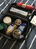 A collection of personal care products placed inside a black travel bag. The items include shower oil, hair wax, deodorant sticks, shaving cream, toothpaste, and a bar of soap. The products are neatly arranged on a fabric with blue and white stripes.
