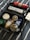 A collection of personal care products placed inside a black travel bag. The items include shower oil, hair wax, deodorant sticks, shaving cream, toothpaste, and a bar of soap. The products are neatly arranged on a fabric with blue and white stripes.
