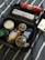A collection of personal care products placed inside a black travel bag. The items include shower oil, hair wax, deodorant sticks, shaving cream, toothpaste, and a bar of soap. The products are neatly arranged on a fabric with blue and white stripes.