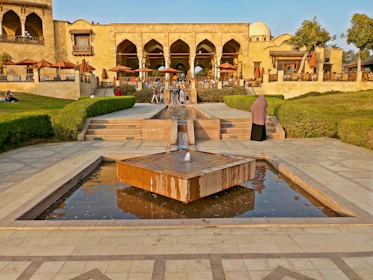 A courtyard features a central water feature in a large public space, surrounded by trimmed hedges and stone pathways. There is a historic-looking building in the background with arched windows and parasols near an outdoor seating area. Several people are present, enjoying the space.