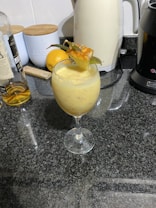 A creamy, yellow beverage served in a clear wine glass, garnished with a piece of pineapple and some pineapple leaves. The glass is placed on a dark granite countertop. In the background, there is a bottle of liquor, a white electric kettle, a black coffee machine, a lemon, a tin labeled 'SUGAR', and a wooden cutting board.