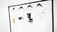 Four white t-shirts hang on black hangers, suspended from a minimalist black clothing rack. The shirts display simple designs, including text and graphic prints in black, and one has an orange rectangular detail. The background is a plain white, emphasizing the clean and modern presentation.