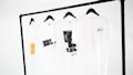 Four white t-shirts hang on black hangers, suspended from a minimalist black clothing rack. The shirts display simple designs, including text and graphic prints in black, and one has an orange rectangular detail. The background is a plain white, emphasizing the clean and modern presentation.