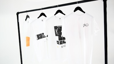 Four white t-shirts hang on black hangers, suspended from a minimalist black clothing rack. The shirts display simple designs, including text and graphic prints in black, and one has an orange rectangular detail. The background is a plain white, emphasizing the clean and modern presentation.