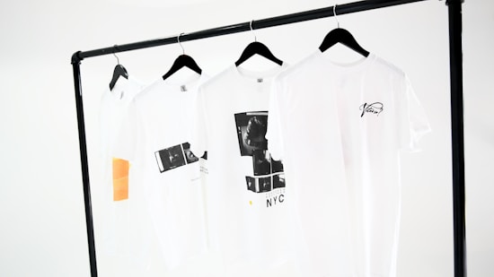 Four white t-shirts hang on black hangers, suspended from a minimalist black clothing rack. The shirts display simple designs, including text and graphic prints in black, and one has an orange rectangular detail. The background is a plain white, emphasizing the clean and modern presentation.