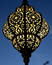 An intricately designed lamp with patterns of swirling and floral motifs. The lamp casts a warm glow against a deep blue evening sky, highlighting its detailed craftsmanship.