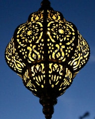An intricately designed lamp with patterns of swirling and floral motifs. The lamp casts a warm glow against a deep blue evening sky, highlighting its detailed craftsmanship.