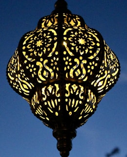 An intricately designed lamp with patterns of swirling and floral motifs. The lamp casts a warm glow against a deep blue evening sky, highlighting its detailed craftsmanship.