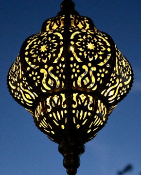 An intricately designed lamp with patterns of swirling and floral motifs. The lamp casts a warm glow against a deep blue evening sky, highlighting its detailed craftsmanship.