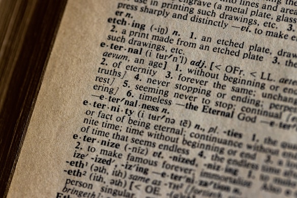 A close-up view of an open dictionary page, focusing on definitions and words in detailed typography. The text is well-aligned, with small, serifed font and some phonetic transcriptions visible.