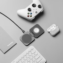 A flat lay of tech accessories on a gray surface, including a white gaming controller, a charging pad, a pair of wireless earbuds in a charging case, a gray notebook labeled 'PocketNote', and a white keyboard.