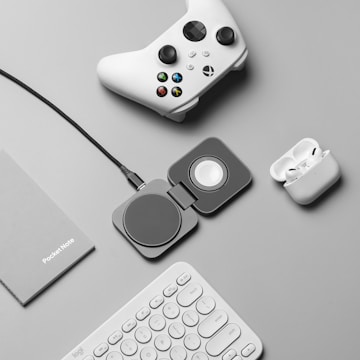 A flat lay of tech accessories on a gray surface, including a white gaming controller, a charging pad, a pair of wireless earbuds in a charging case, a gray notebook labeled 'PocketNote', and a white keyboard.
