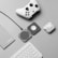 A flat lay of tech accessories on a gray surface, including a white gaming controller, a charging pad, a pair of wireless earbuds in a charging case, a gray notebook labeled 'PocketNote', and a white keyboard.