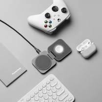 A flat lay of tech accessories on a gray surface, including a white gaming controller, a charging pad, a pair of wireless earbuds in a charging case, a gray notebook labeled 'PocketNote', and a white keyboard.