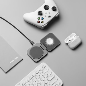 A flat lay of tech accessories on a gray surface, including a white gaming controller, a charging pad, a pair of wireless earbuds in a charging case, a gray notebook labeled 'PocketNote', and a white keyboard.