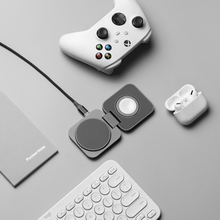 A flat lay of tech accessories on a gray surface, including a white gaming controller, a charging pad, a pair of wireless earbuds in a charging case, a gray notebook labeled 'PocketNote', and a white keyboard.