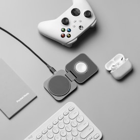 A flat lay of tech accessories on a gray surface, including a white gaming controller, a charging pad, a pair of wireless earbuds in a charging case, a gray notebook labeled 'PocketNote', and a white keyboard.