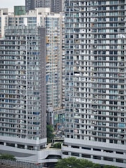 Tall residential buildings dominate the urban landscape with numerous windows and balconies. The structures are tightly packed, showcasing a densely populated area with a modern architectural style. Green spaces are visible at the base, providing a contrast to the concrete structures.