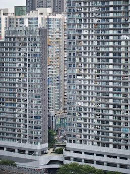 Tall residential buildings dominate the urban landscape with numerous windows and balconies. The structures are tightly packed, showcasing a densely populated area with a modern architectural style. Green spaces are visible at the base, providing a contrast to the concrete structures.