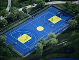 A vibrant outdoor basketball court with a blue surface and yellow accents, surrounded by lush green trees and neatly manicured lawns. Two people are present on the court, engaged in playing basketball or practice.