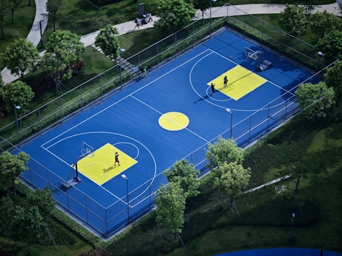 A vibrant outdoor basketball court with a blue surface and yellow accents, surrounded by lush green trees and neatly manicured lawns. Two people are present on the court, engaged in playing basketball or practice.