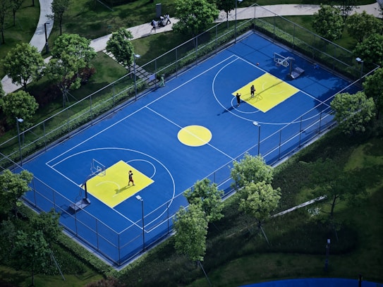 A vibrant outdoor basketball court with a blue surface and yellow accents, surrounded by lush green trees and neatly manicured lawns. Two people are present on the court, engaged in playing basketball or practice.