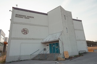 A large, white industrial-style building with a rectangular shape and corrugated metal exterior. There are faded letters reading 'Burnaby North Secondary School' and a circular emblem in red on the facade. The structure features a small overhang above the entrance, which has teal-colored double doors, accessible via a short set of concrete steps with metal railings. A few utility structures and equipment are scattered around, with signs of some wear and weathering on the walls.